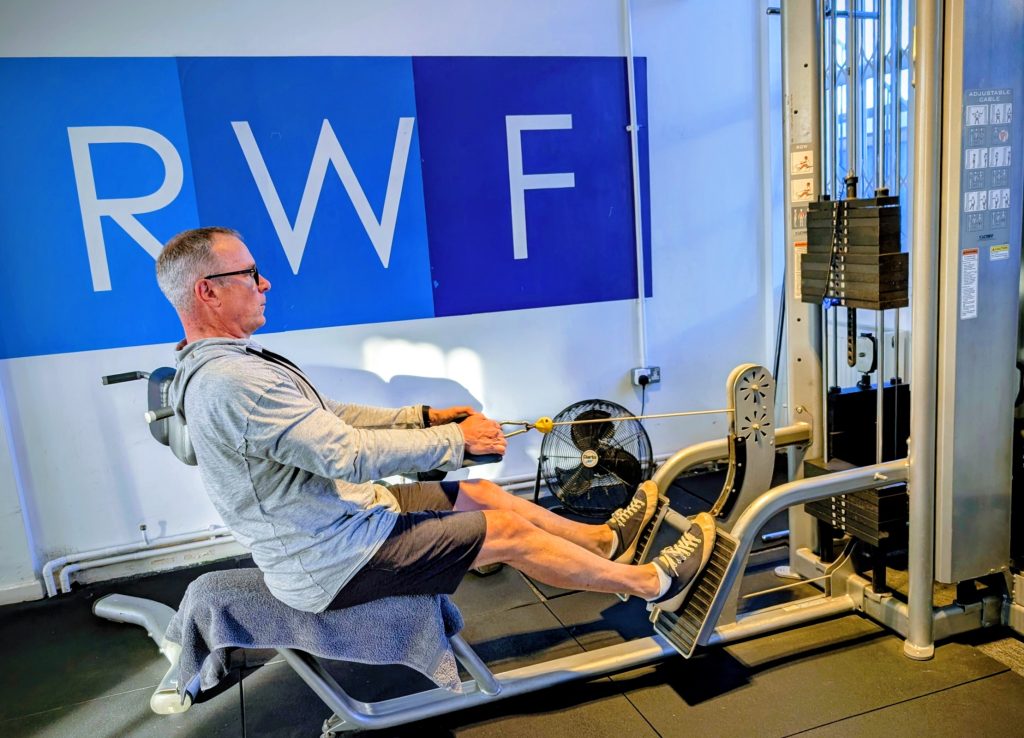 Man seated in a cable-resistance rehab machine, pulling on handles with legs supported, in a room with a blue wall mural behind him.