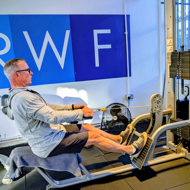 Man seated in a cable-resistance rehab machine, pulling on handles with legs supported, in a room with a blue wall mural behind him.
