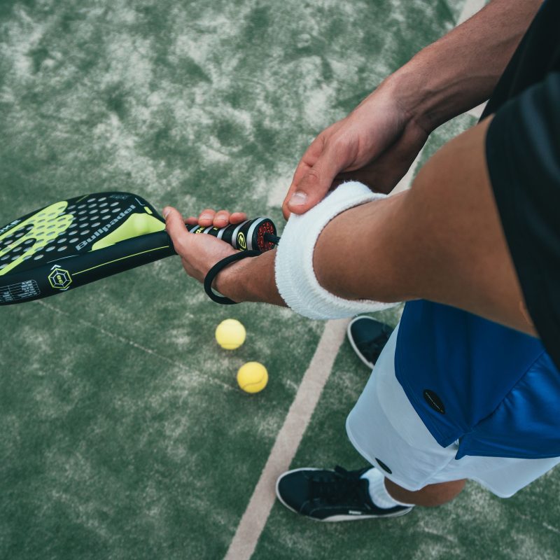 Person on a tennis/padel court tucks a white towel around their ankle while holding a black-yellow perforated padel racket; several tennis balls on the ground nearby.