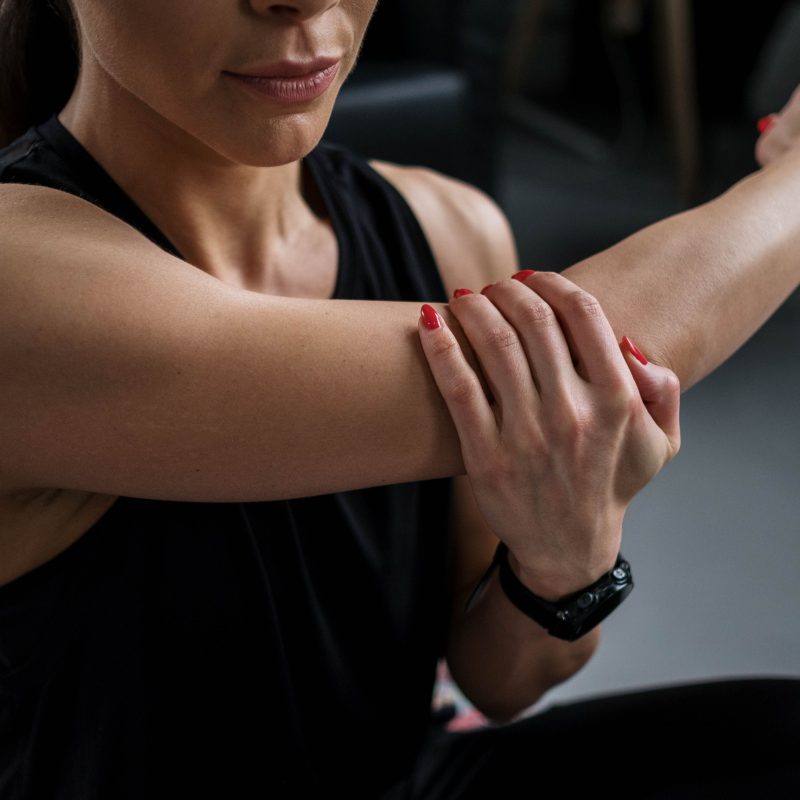 Person in a black sleeveless top stretches their left arm across the chest, aided by the right hand, with red nails visible.