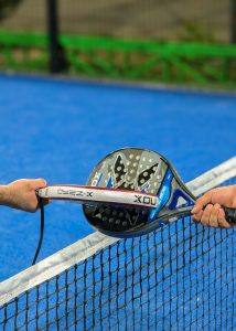 pexels-zokirkodirov-37978261 | Real World Fitness - Nottingham Gym, Personal Training and Sports Massage Two hands pass a perforated paddle/board over a net on a bright blue court, suggesting a paddle sport exchange.