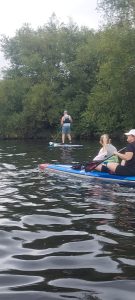 1000036484 | Real World Fitness - Nottingham Gym, Personal Training and Sports Massage Man standing on a stand-up paddleboard paddling on a calm river with a tree-lined bank behind him.