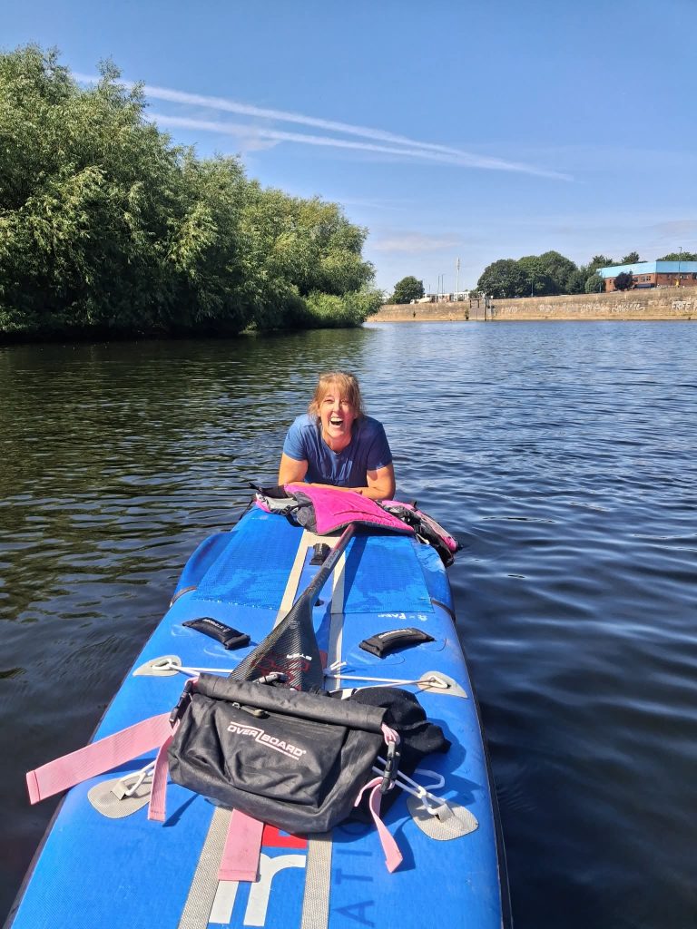 Smiling woman on a blue stand-up paddleboard paddling on a calm river with trees on the left and a brick wall and buildings in the background.
