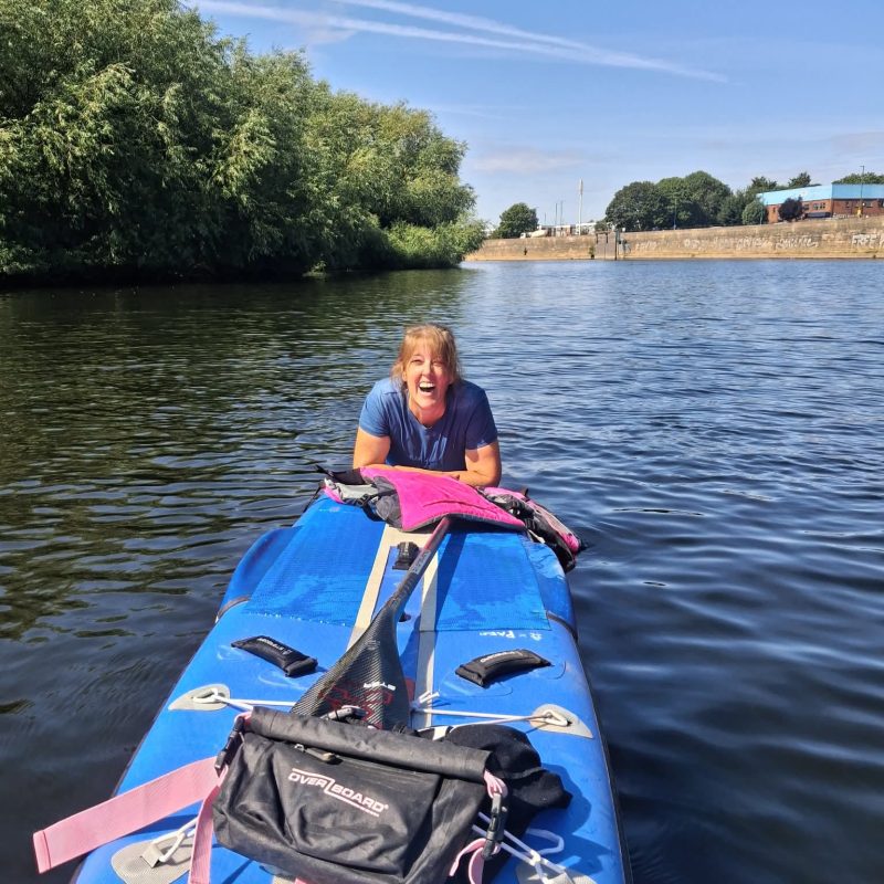 Smiling woman on a blue stand-up paddleboard paddling on a calm river with trees on the left and a brick wall and buildings in the background.